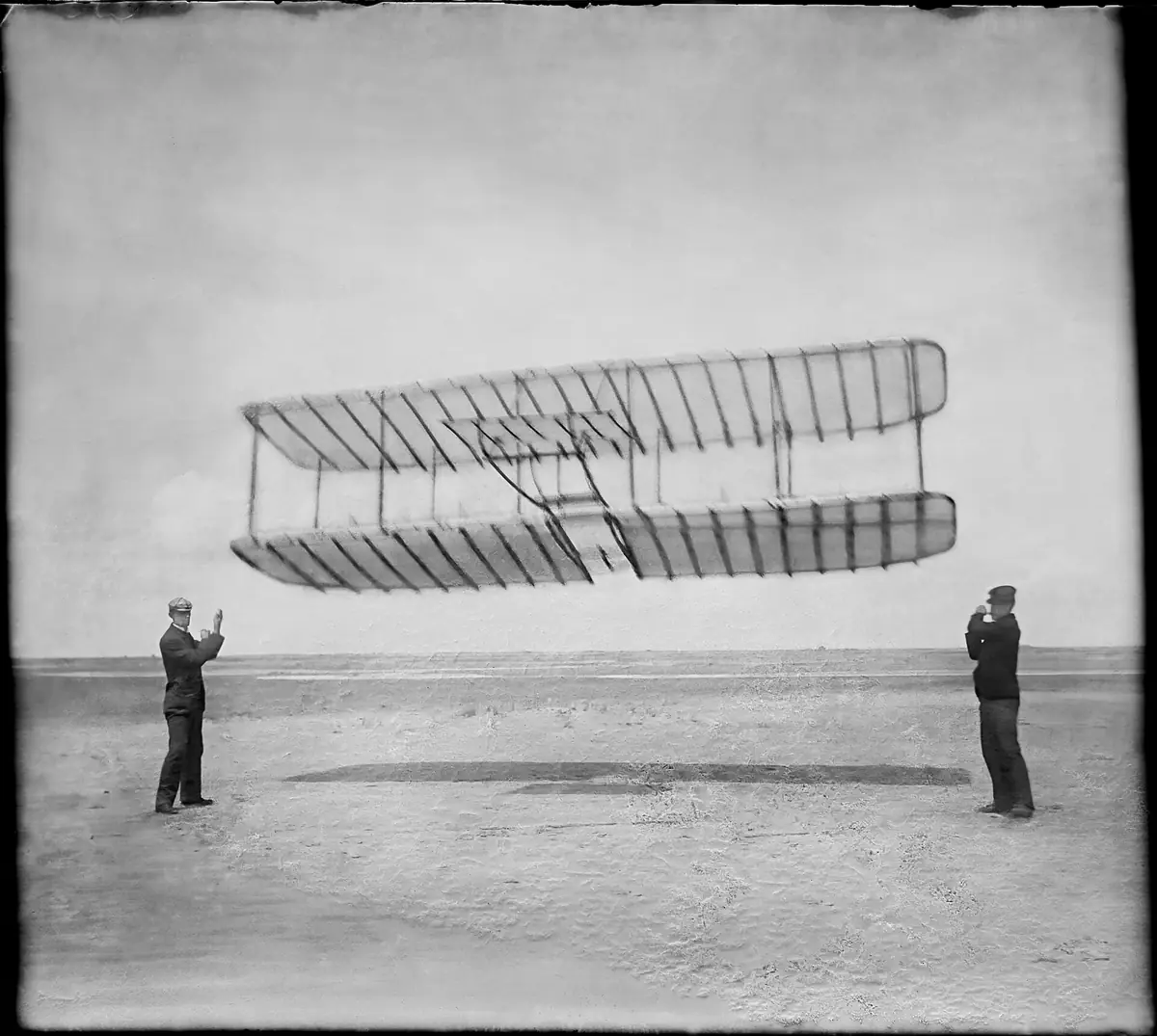 Wilbur and Orville Wright holding the lines of their 1901 glider as it flies like a kite above the sand.